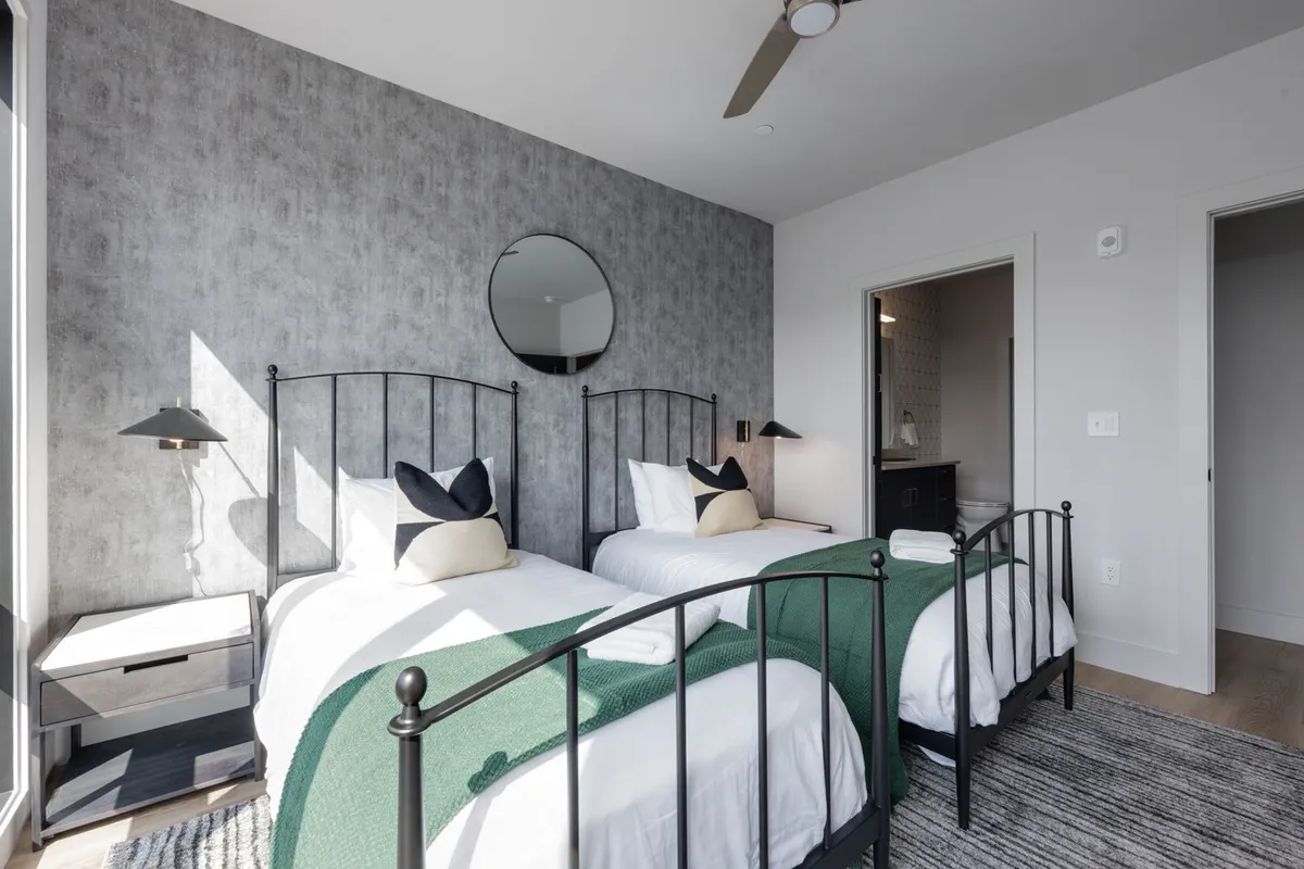 A welcoming and cozy twin bedroom, with two black metal-framed beds adorned with green throws. The room is complemented by a round mirror and soft lighting, inviting relaxation and togetherness.