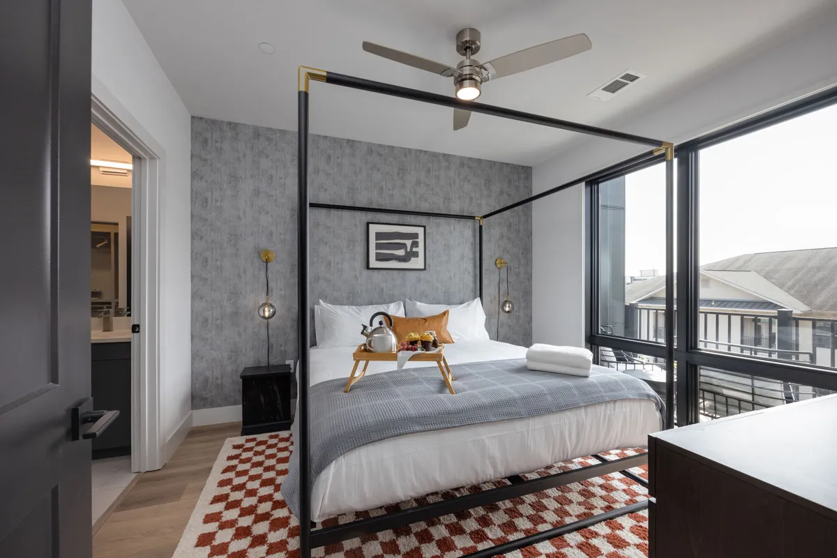 A serene, modern bedroom with a four-poster king bed dressed in stylish bedding. Natural light streams through a large window, illuminating the room and its decorative rug, wall art, and a smart TV visible for entertainment.