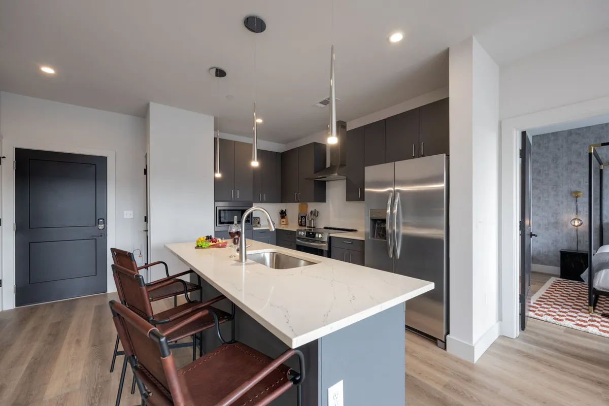 This image captures a modern kitchen with a large island serving as a breakfast bar, surrounded by stylish bar stools. The kitchen features gleaming stainless steel appliances and elegant light fixtures overhead. The space is shown in use for both cooking and entertaining, evoking a lively, welcoming atmosphere.