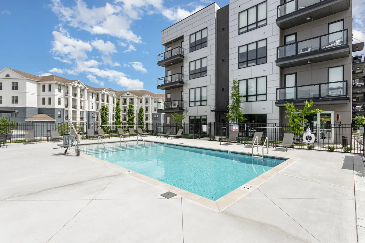 A tranquil exterior view of a resort-style swimming pool surrounded by green trees and landscaping, with the balconies of the apartment building overlooking the water. The pool area features spaces for sunbathing, social gatherings, and relaxing by the water, evoking a resort-like atmosphere.