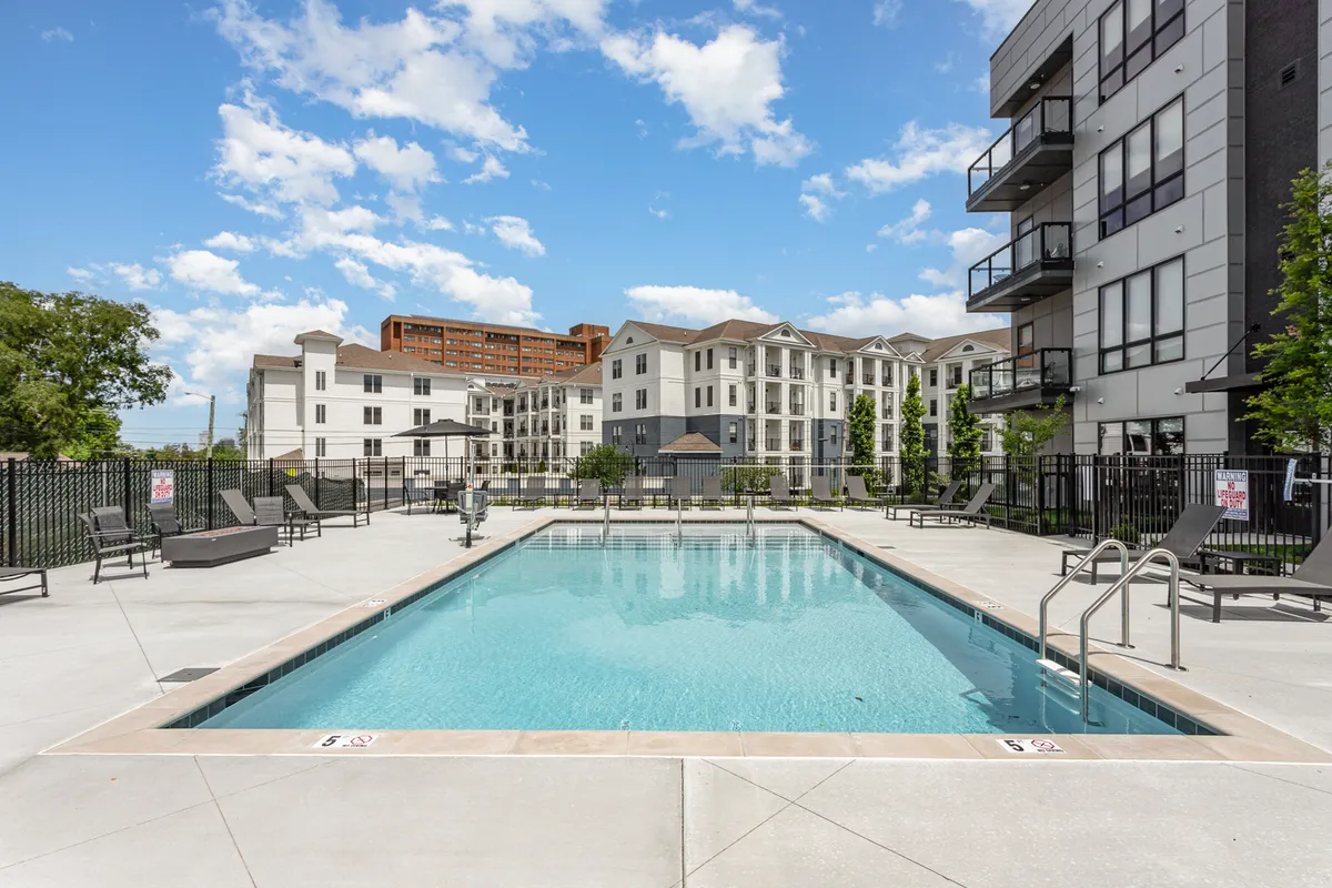 An inviting view of a swimming pool with lounge chairs and a landscaped area, set against the backdrop of residential buildings. The scene suggests opportunities for sunbathing, relaxation, and social gatherings by the pool.